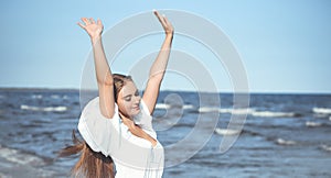 Happy smiling beautiful woman on the ocean beach standing in a white summer dress, raising hands