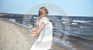 Happy smiling beautiful woman is on the ocean beach in a white summer dress, open arms