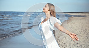 Happy smiling beautiful woman is on the ocean beach in a white summer dress, open arms