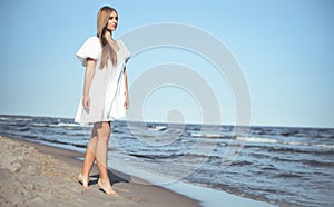 Happy smiling beautiful woman is walking on the ocean beach in a white summer dress