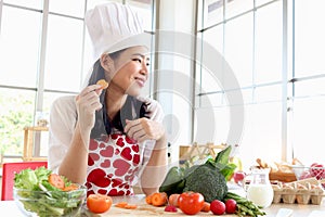 Happy smiling beautiful young Asian woman in red heart apron and chef hat holds and eats sliced carrot while sits behind kitchen