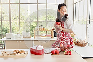 Happy smiling beautiful young Asian woman in red heart apron holds red coffee mug while stands behind kitchen counter. Girl