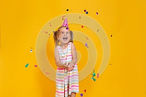 Happy smiling birthday child girl in pink cup surrounded by flying confetti on colored yellow background . Celebration