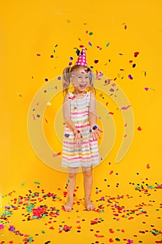 Happy smiling birthday child girl in pink cup surrounded by flying confetti on colored yellow background . Celebration