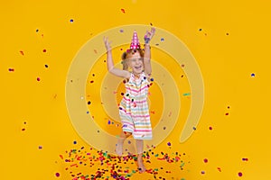 Happy smiling birthday child girl in pink cup surrounded by flying confetti jumping on colored yellow background