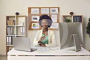 Happy smiling black woman sitting at office desk with laptop and desktop computer