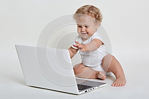 Happy smiling blond curly haired child sitting barefoot on floor with laptop computer and pulls his hands to screen, has positive