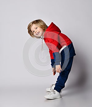 Happy smiling blond kid boy in blue and red hoodie and pants poses sideways bending over reaching his hands to toes