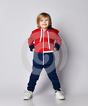 Happy smiling blond kid boy in red and blue sportwear and white sneakers poses holding hands at his waist photo