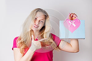 Happy smiling blonde woman pointing finger at blue gift bag with pink heart on white background