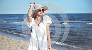 Happy smiling blonde woman is posing on the ocean beach with sunglasses and a hat