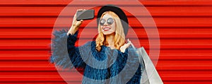 Happy smiling blonde woman taking selfie picture by smartphone with shopping bags wearing blue faux fur coat, black round hat