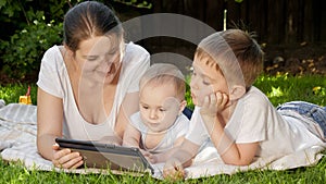 Happy smiling boy with baby brother and mother lying on grass in park and browsing internet on tablet computer