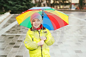 Happy smiling boy enjoying rain in city. Kid has colorful rainbow umbrella. Child wears waterproof warm jacket. Autumn rainy