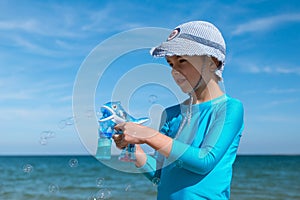 Happy smiling boy the European in a blue UF protective t-shirt and red shorts on the beach by the blue sea starts up soap bubbles