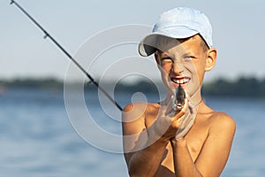 Happy smiling boy holding taken freshwater fish in hands against rod and river background