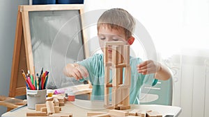 Happy smiling boy looking at toy wooden blocks tower he built on desk at school classroom. Concept of smart children and