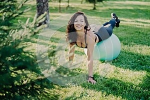 Happy smiling brunette woman has yoga exercises with fitness ball poses at park with green grass, has cheerful expression, stays