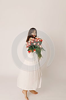 Happy smiling brunette woman in white dress holding tulips in her hands and looking at the camera, 8 march