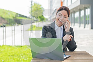 Happy smiling business woman talking on her mobile and browsing on a laptop while sitting at a table in a cafe on a