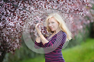 Happy smiling Caucasian blond woman with long hair in purple fedora hat near blossoming plum cherry tree, enjoys the blossom