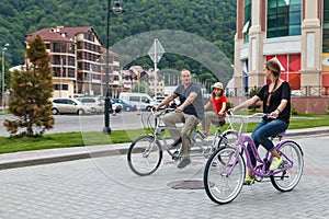 Happy smiling Caucasian family of father, mother and daughter have outdoor bicycle ride at summer on hotel buildings background.