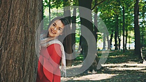 Happy smiling caucasian girl is showing up from tree caudex, isolated, on blurred park background. Close up. Copy space.