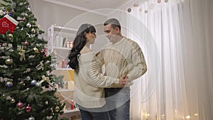 Happy smiling Caucasian husband and pregnant wife dancing in slow motion on Christmas eve at home. Cheerful loving man