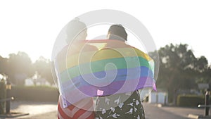 Happy smiling Caucasian lesbian couple walking in sunrays with rainbow flag talking. Back view of positive LGBT women