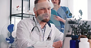 Happy smiling Caucasian male doctor wearing lab coat, stethoscope talks to remote client online via laptop webcam video.