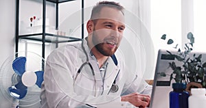 Happy smiling Caucasian male doctor wearing lab coat, stethoscope talks to remote client online via laptop webcam video.