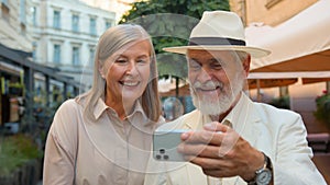 Happy smiling Caucasian senior elderly couple walking taking selfie laughing talking gadget mobile phone together city