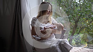 Happy smiling Caucasian woman sitting on windowsill shaking newborn baby in slow motion looking out the window. Portrait