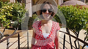 Happy smiling caucasian woman walking towards the camera wearing red dress and sunglasses in the hotel palm tree garden. Closeup s