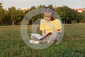 happy smiling cheerful girl 7 years old reads book, sits on grass in park and enjoys in summer day