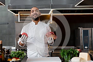 Happy smiling chef prepares meat dish with various vegetables in the kitchen. In one hand the man holds vegetables, in the other a