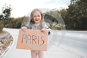 Happy smiling child girl 3-4 year old hold paper craft Paris handwritten sign hitch hiking on road outdoors. Happiness.