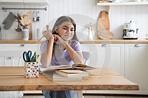 Happy smiling child teenage girl studying alone at home, reading textbook and doing homework