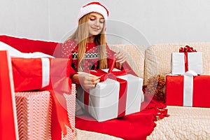 Happy smiling child wearing Santa Claus hat opening Christmas present while sitting on sofa at home  Merry Christmas and Holidays