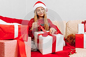 Happy smiling child wearing Santa Claus hat opening Christmas present while sitting on sofa at home  Merry Christmas and Holidays