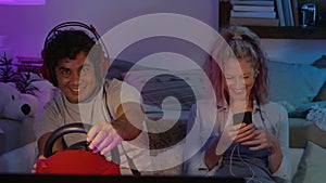 Happy smiling couple at home in evening, young wife uses smartphone and her husband plays computer video games, fun in living room