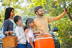 Happy smiling couple with kids taking selfie on mobile phone during summer camp picnic in front of car - concept of