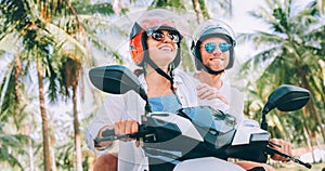 Happy smiling couple travelers riding motorbike scooter in safety helmets during tropical vacation under palm trees on Ko Samui