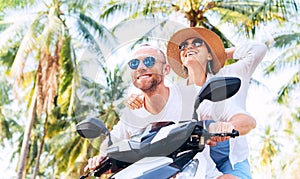 Happy smiling couple travelers riding motorbike during their tropical vacation under palm trees