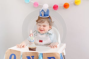 Happy smiling cute Caucasian baby boy in blue crown celebrating his first birthday at home. Child kid toddler sitting in high