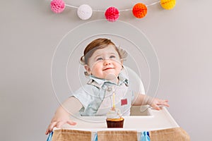 Happy smiling cute Caucasian baby boy celebrating his first birthday at home. Child kid toddler sitting in high chair eating tasty