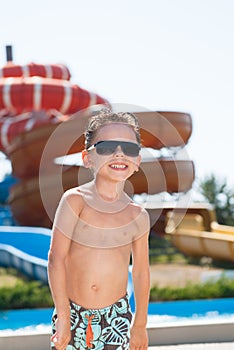 Happy smiling cute liitle boy in shorts and sunglasses in water park with water slides in summer