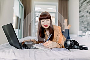 Happy smiling dark haired woman lefthander using laptop and making notes in her diary notepad, while lying on bed in the