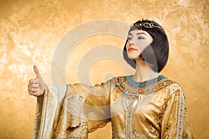 Happy smiling Egyptian woman like Cleopatra with thumbs up gesture, on golden background