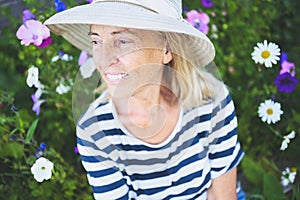 Happy smiling elderly senior woman having fun posing in summer garden with flowers in straw hat. Farming, gardening, agriculture,